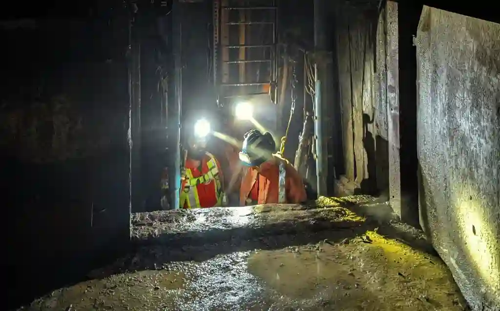 Two workers in helmets and safety harnesses inspecting a mine shaft beside a ladder and structural supports.